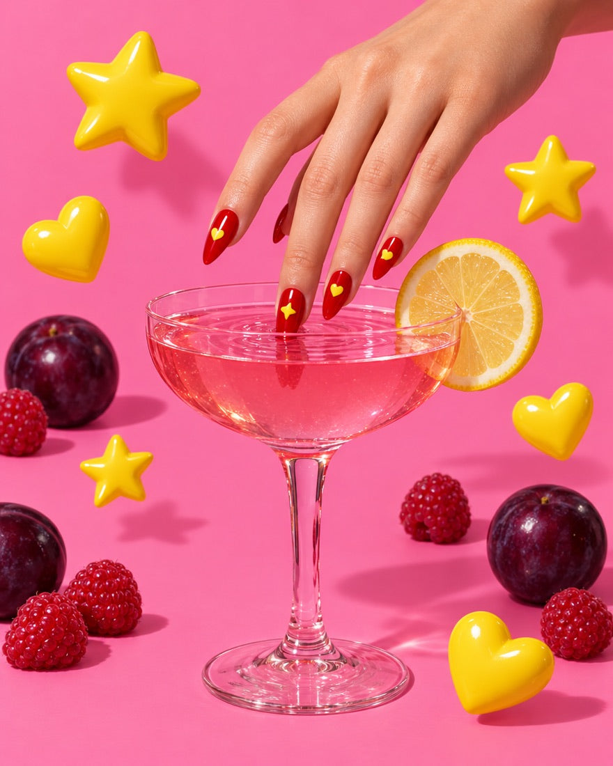 Mano con las uñas rojas con pegatinas de corazón y estrella, mojando una uña en una copa de cóctel rosa, entre corazones y estrellas amarillos